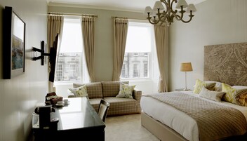 A hotel room with a bed, a desk, a chair, a lamp, and a view of buildings through the windows.