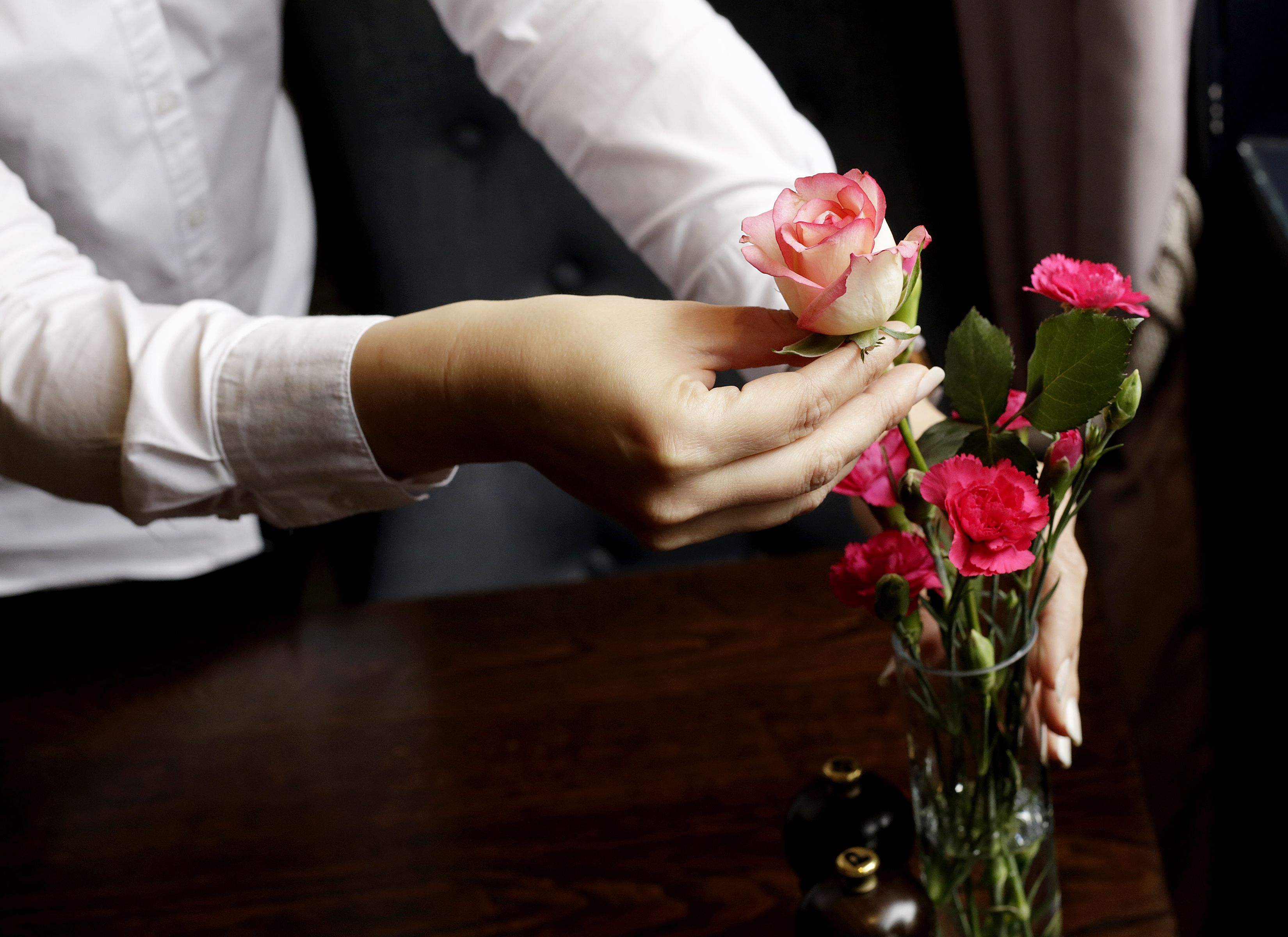A hand holding a rose, with a vase of flowers on a table.