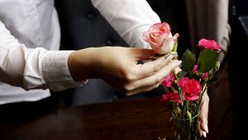 A hand holding a rose, with a vase of flowers on a table.