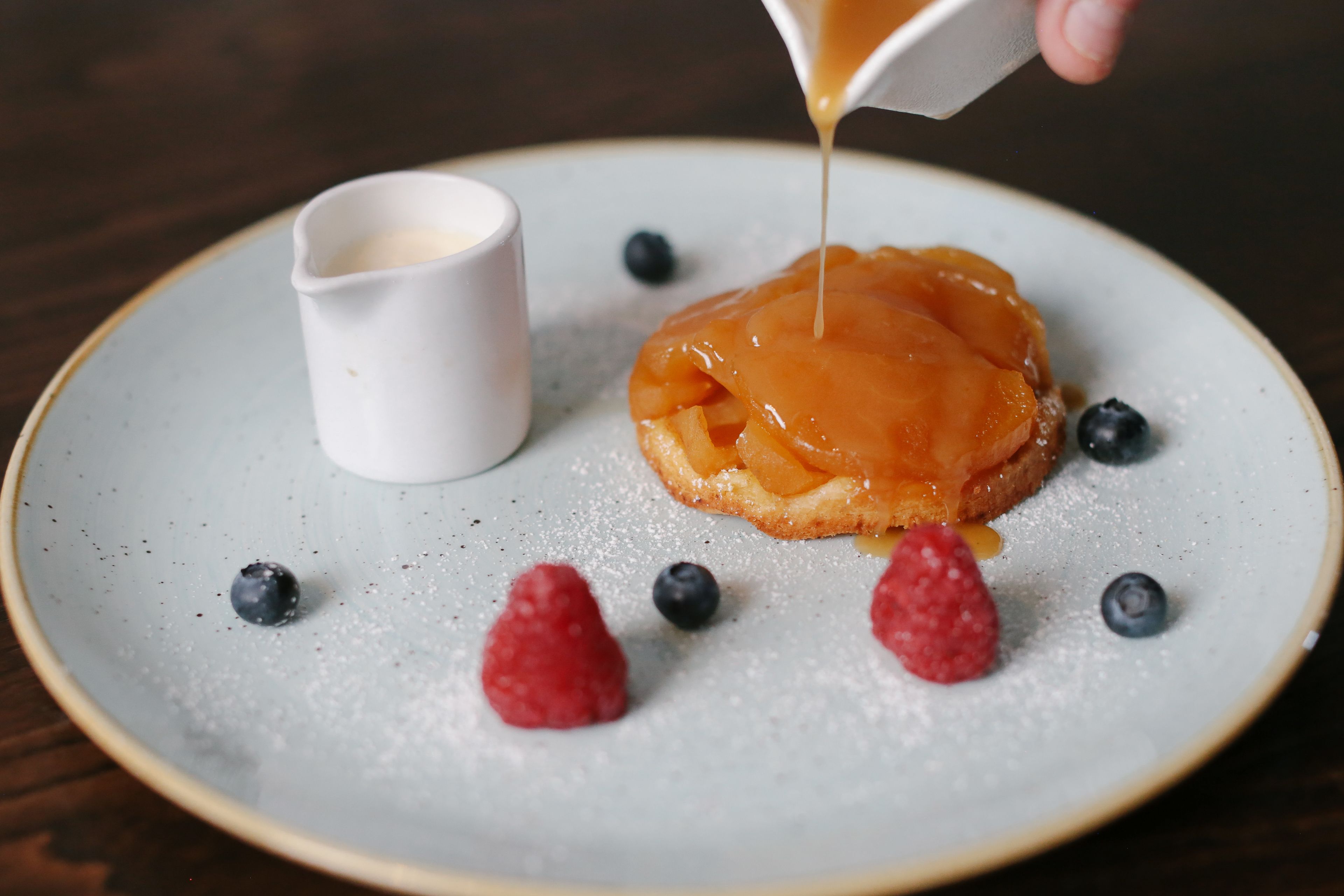 A dessert plate with a caramel apple, blueberries, and raspberries, and a small cup of cream.