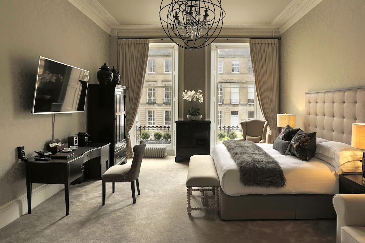 Une chambre d’hôtel comprenant un lit, une table de chevet, une lampe, une chaise, un bureau, une télévision, une commode et une vue sur les bâtiments à travers de grandes fenêtres.
