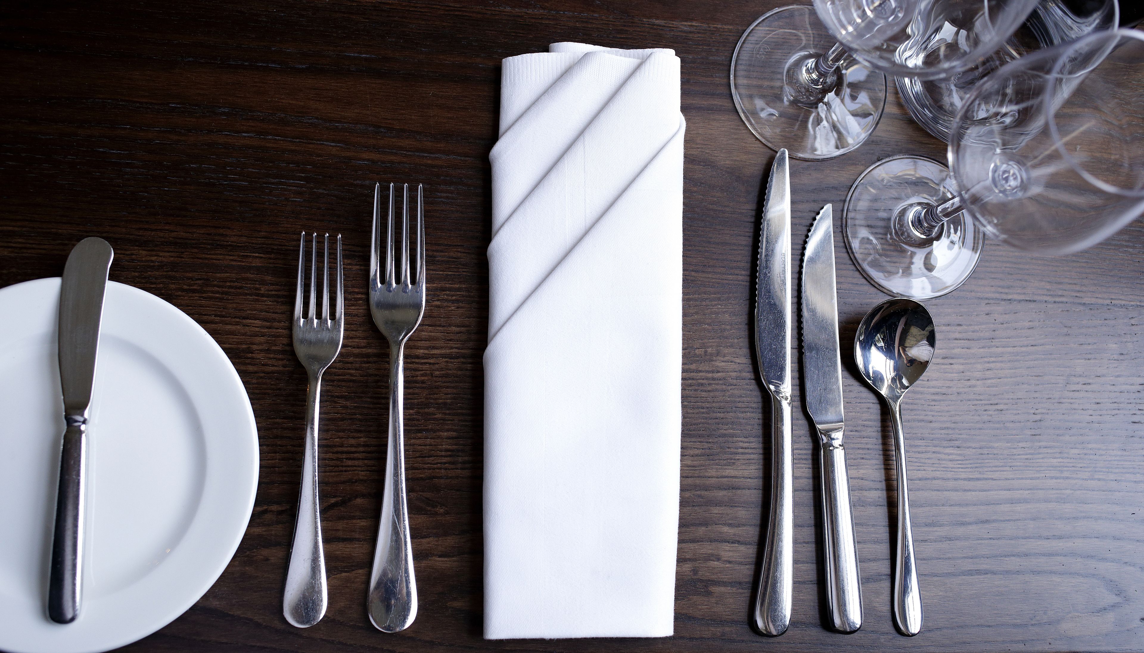 A neatly arranged table setting with a white napkin, silverware, and wine glasses on a wooden table.