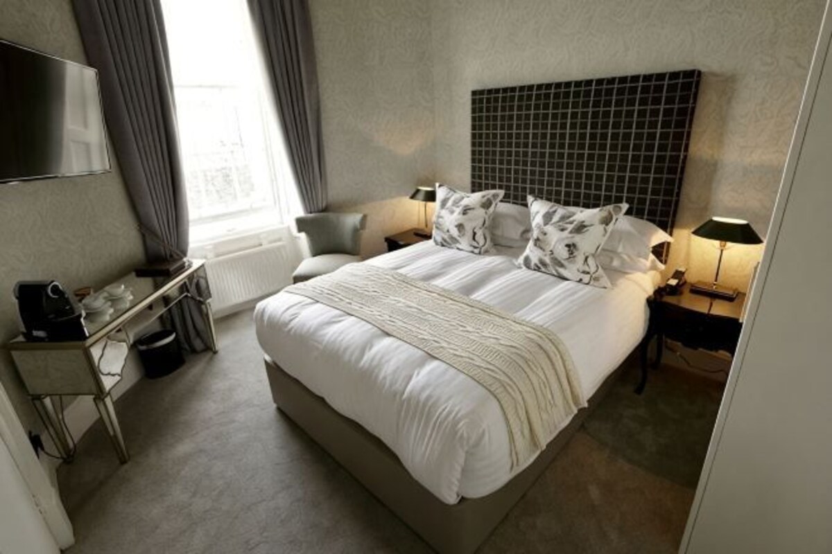 A hotel room with a large bed, a desk with a chair, a TV mounted on the wall, and a window with curtains.