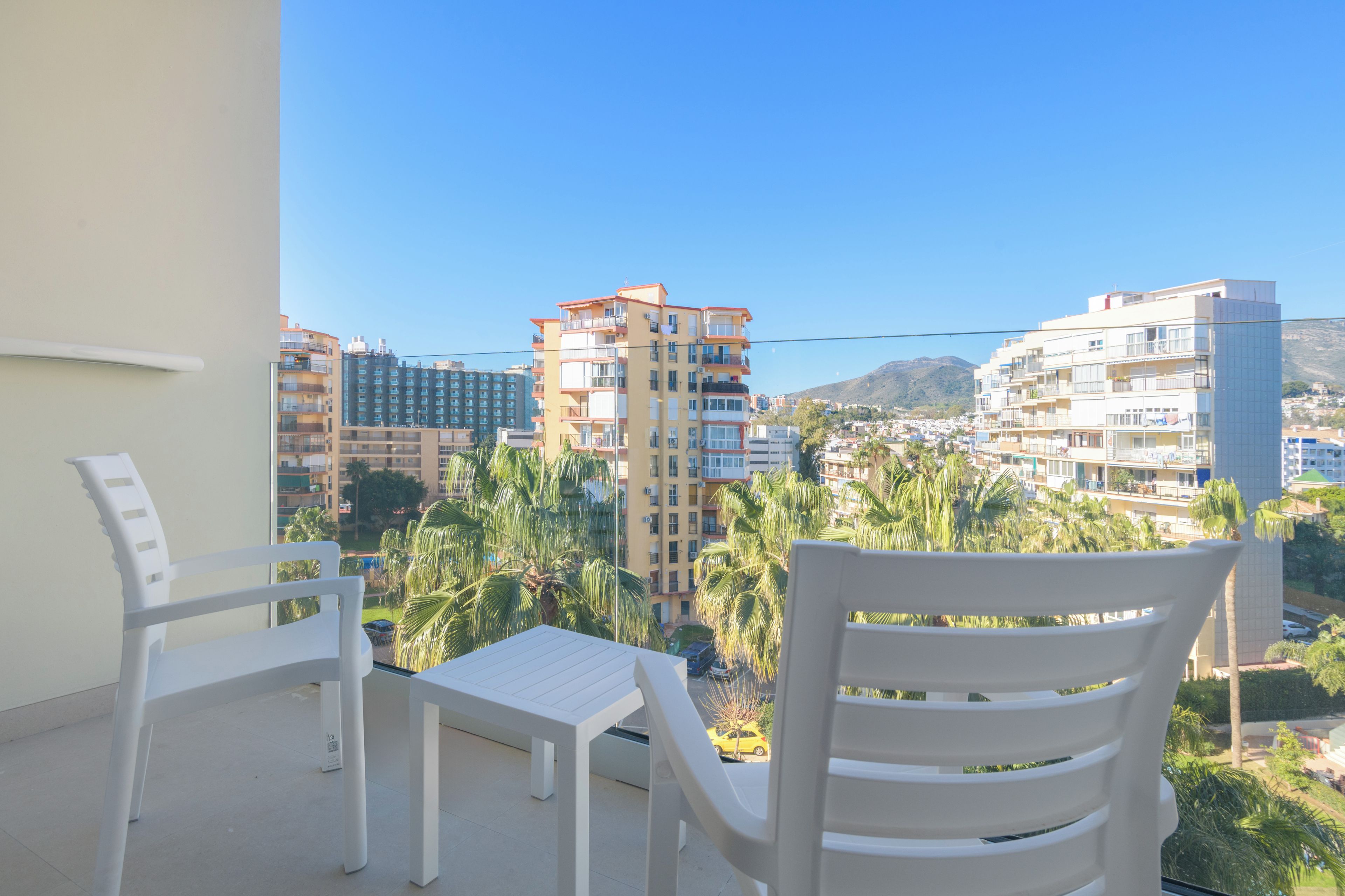 A balcony with white chairs and a table, overlooking a cityscape with multi-story buildings and palm trees.