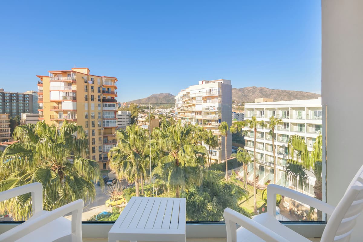A balcony with white chairs and a table, overlooking a cityscape with palm trees and buildings.