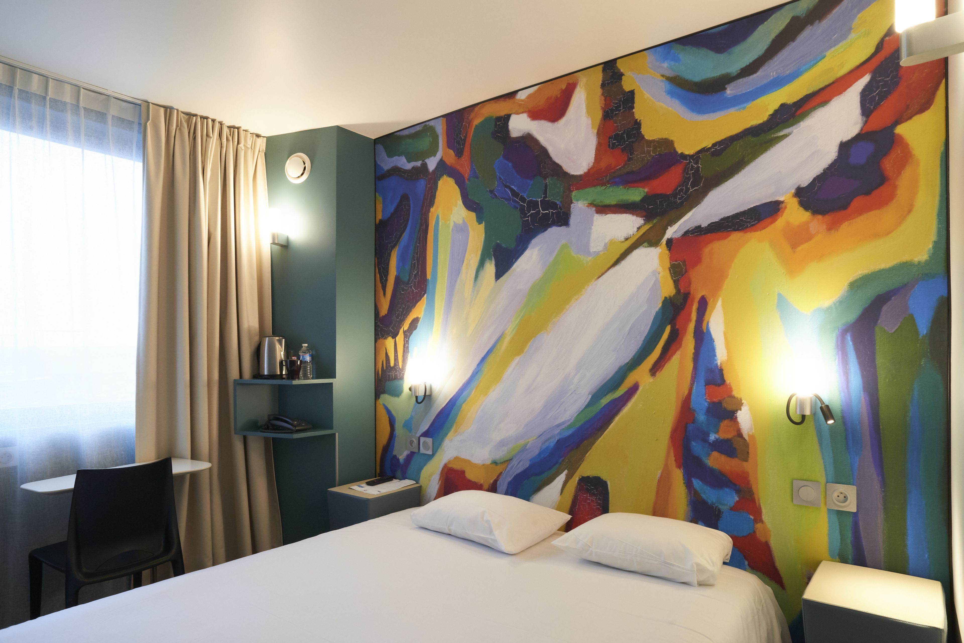 A hotel room with a bed, a desk, and a chair. There is a colorful abstract painting on the wall.