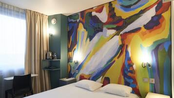A hotel room with a bed, a desk, and a chair. There is a colorful abstract painting on the wall.