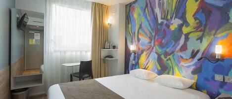 A hotel room with a bed, a desk, a chair, a mirror, a colorful wall, and a nightstand with a lamp.