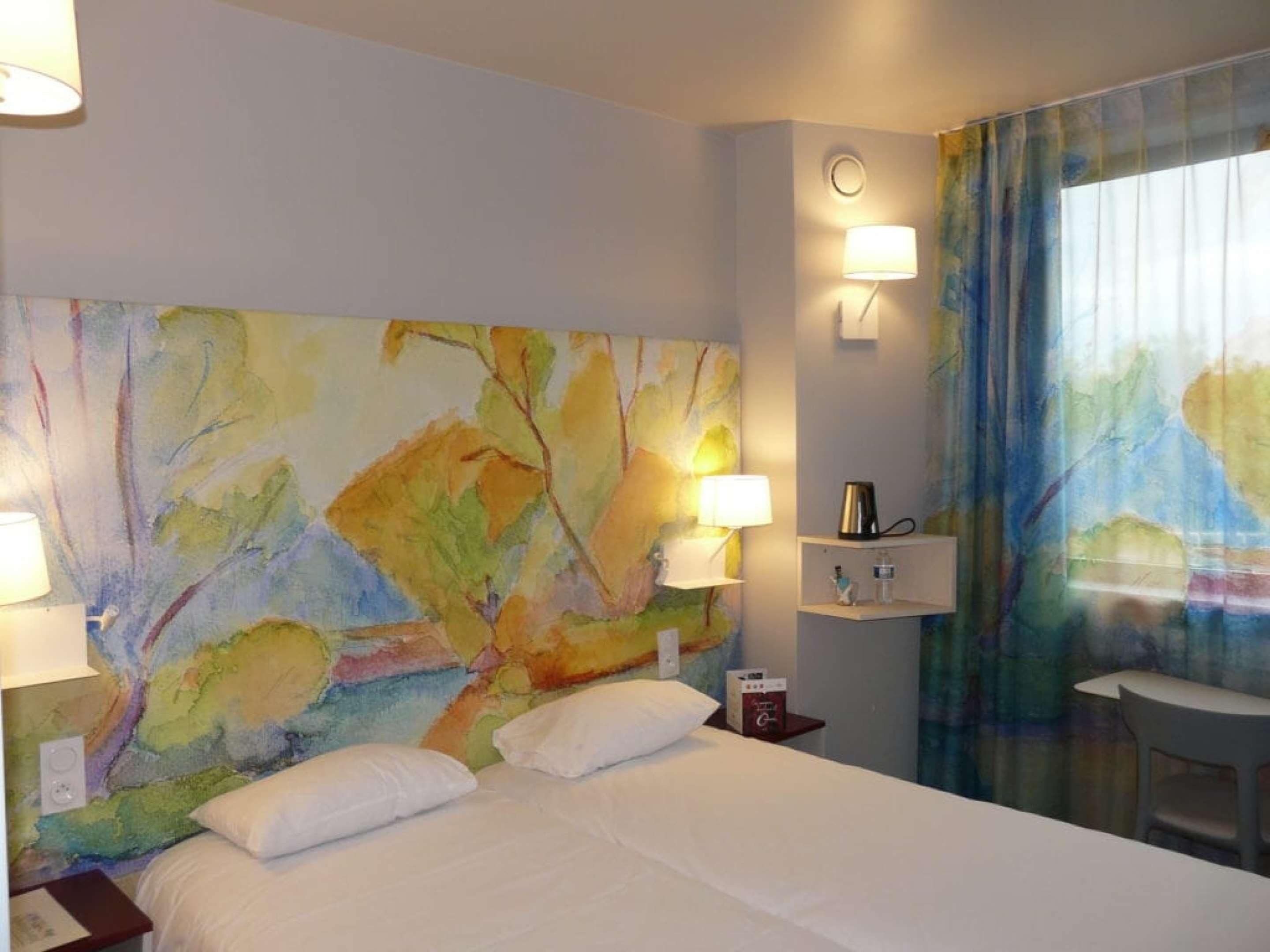 A hotel room with a large abstract painting on the wall, a bed with white linens, two bedside lamps, a small table with a cup and a book, and a window with curtains.