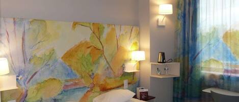 A hotel room with a large abstract painting on the wall, a bed with white linens, two bedside lamps, a small table with a cup and a book, and a window with curtains.
