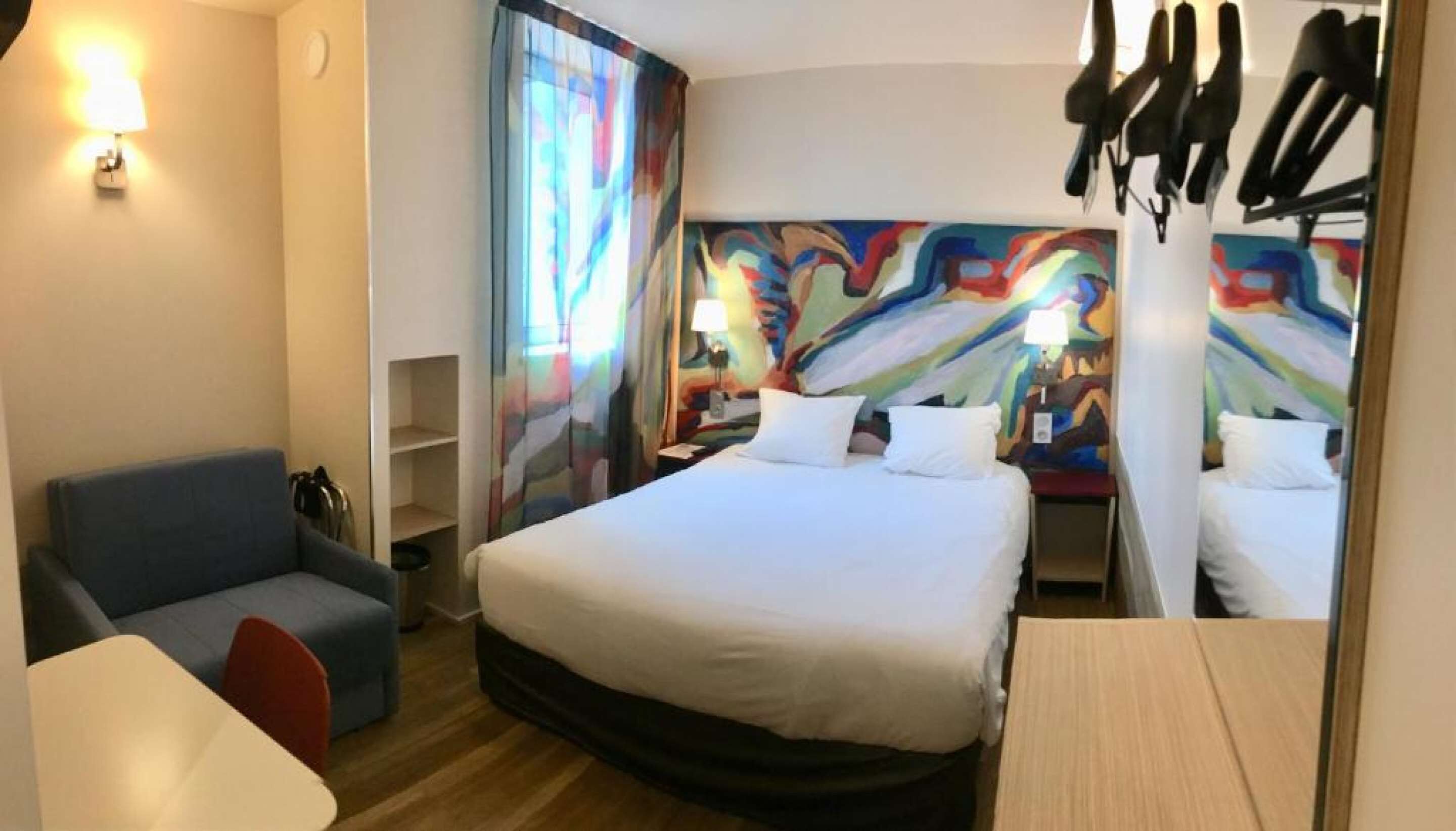 Habitación de hotel con cama, escritorio, silla, lámpara y un cuadro abstracto colorido en la pared.