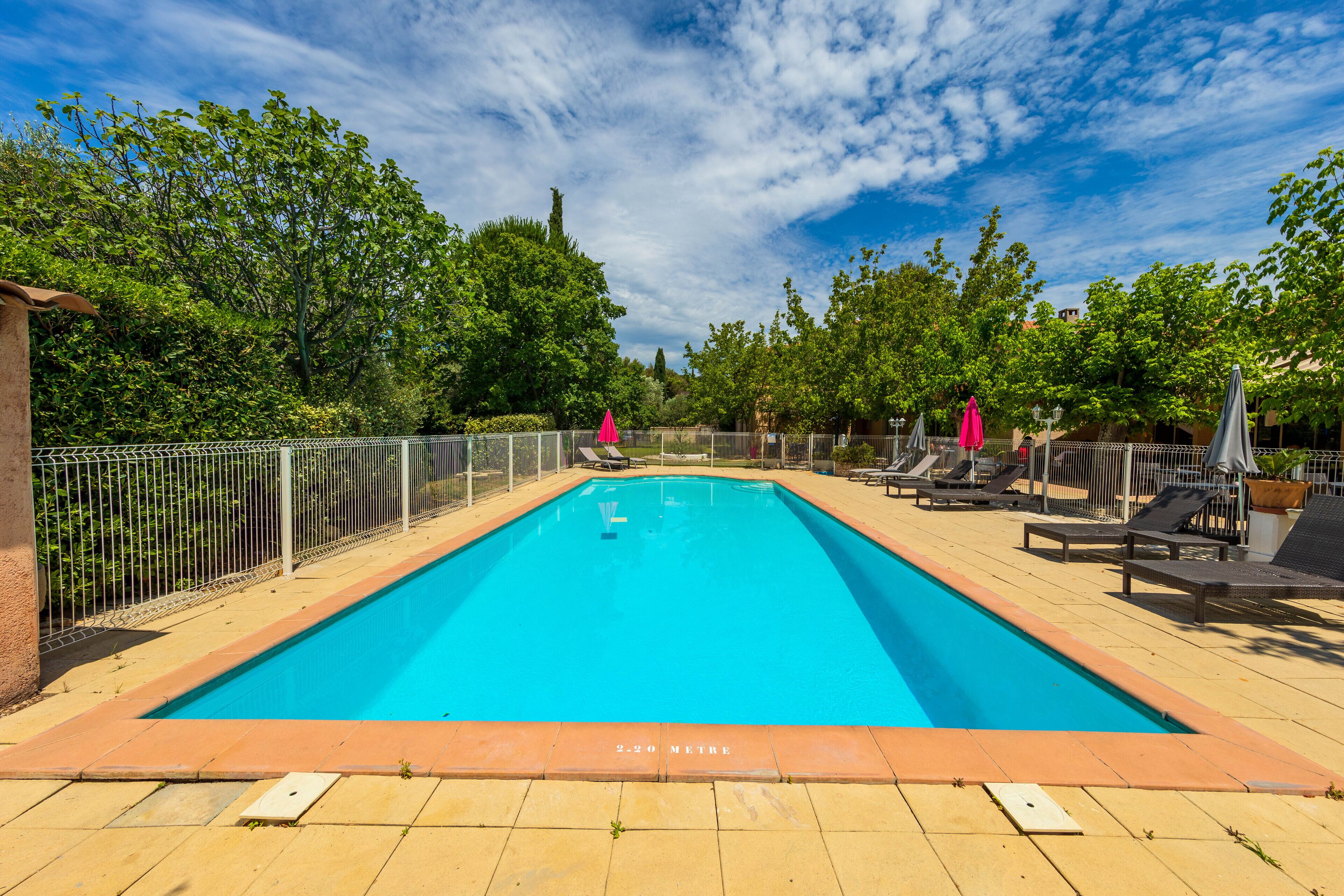 Une piscine rectangulaire, dotée d’une terrasse carrelée, entourée d’une clôture en métal et ombragée par des parasols.
