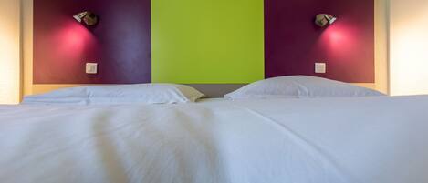 Un lit double avec du linge de lit blanc, une tête de lit verte et violette, et deux lampes fixées au mur.