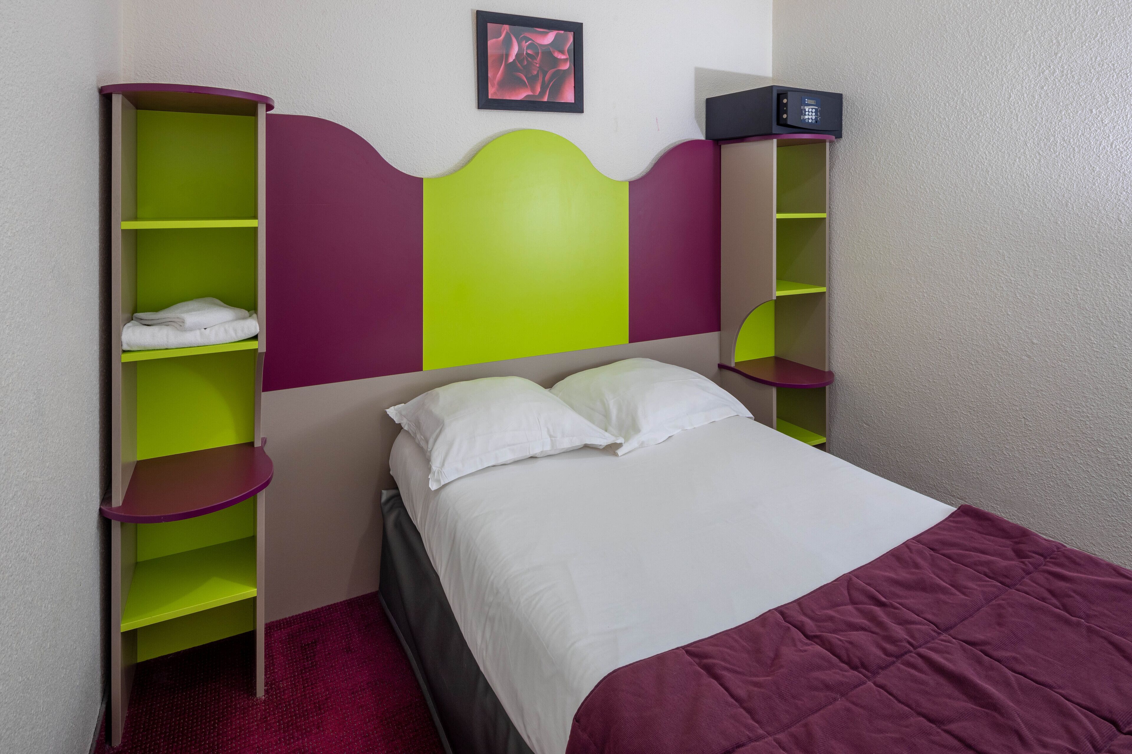 Quarto com cama, cabeceira verde e roxa, edredom branco e prateleira com micro-ondas.