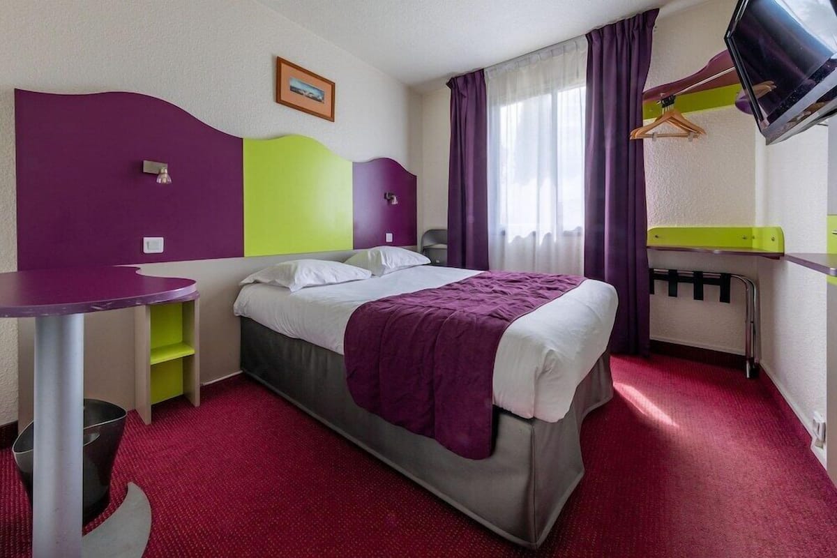 A hotel room with a bed, a desk, a chair, a TV, and a window with curtains.