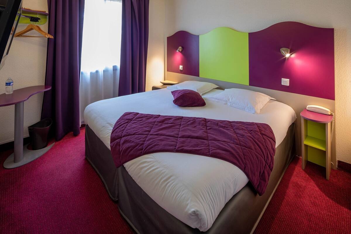 A hotel room with a bed, a bedside table, a nightstand, a hanging coat rack, and a window with curtains.