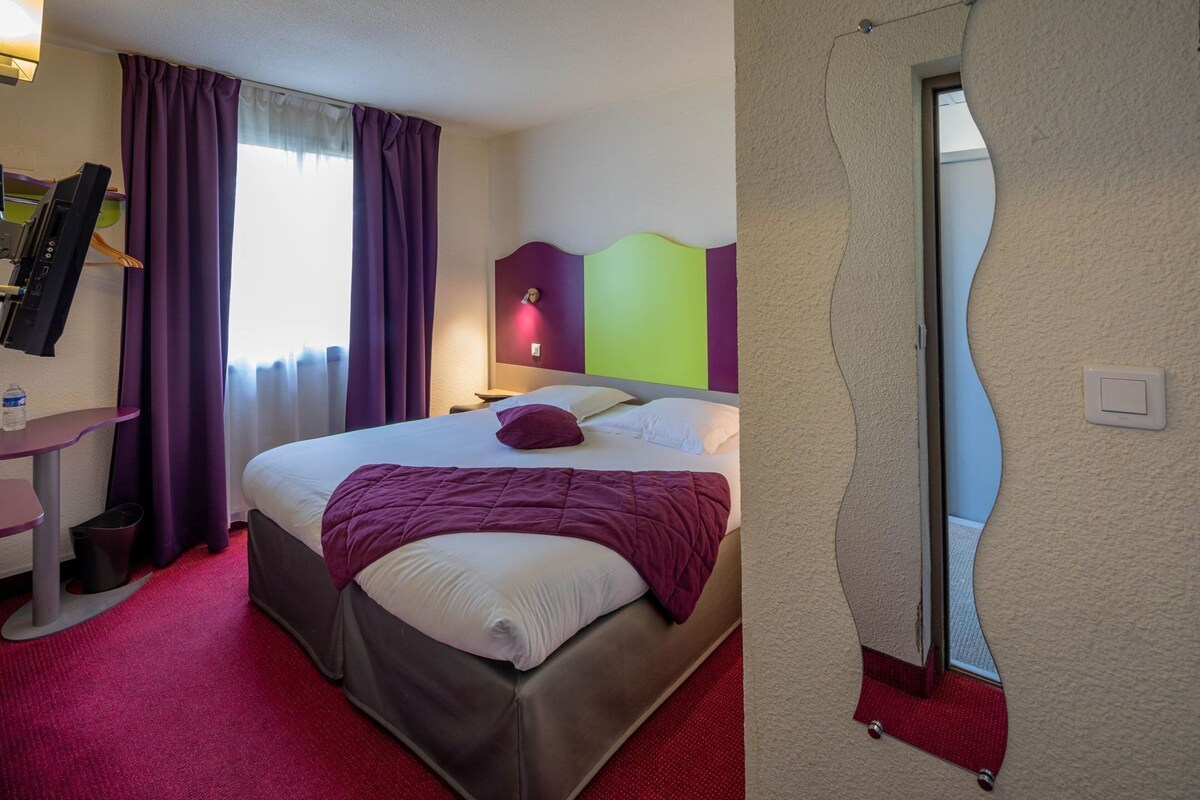Une chambre d’hôtel avec un lit, un bureau, un miroir et une fenêtre avec des rideaux.