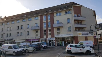 Um edifício de vários andares, com um rés-de-chão que inclui uma salão de cabeleireiro e uma loja de cosméticos. Vários carros estão estacionados em frente, e uma placa de hotel é visível no lado direito.