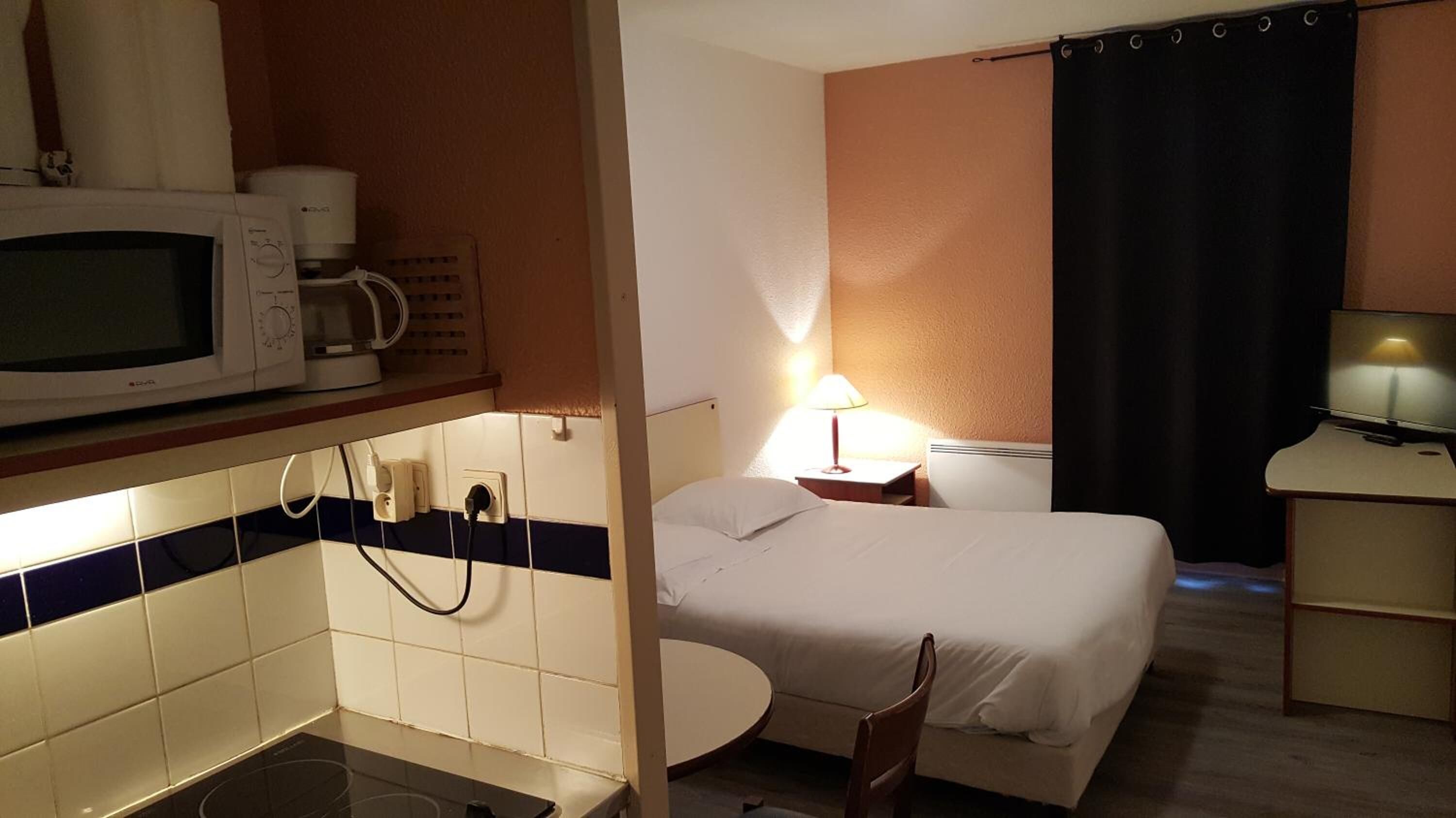 Hotelkamer met een bed, nachtkastje met lamp, televisie, magnetron en een kleine kitchenette met fornuis.