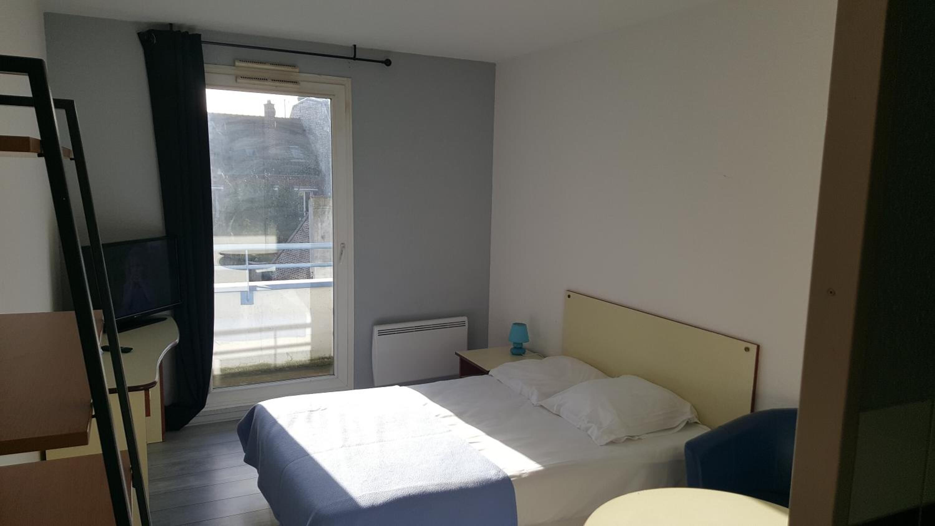 A hotel room with a bed, a desk, a chair, a radiator, and a window with curtains.