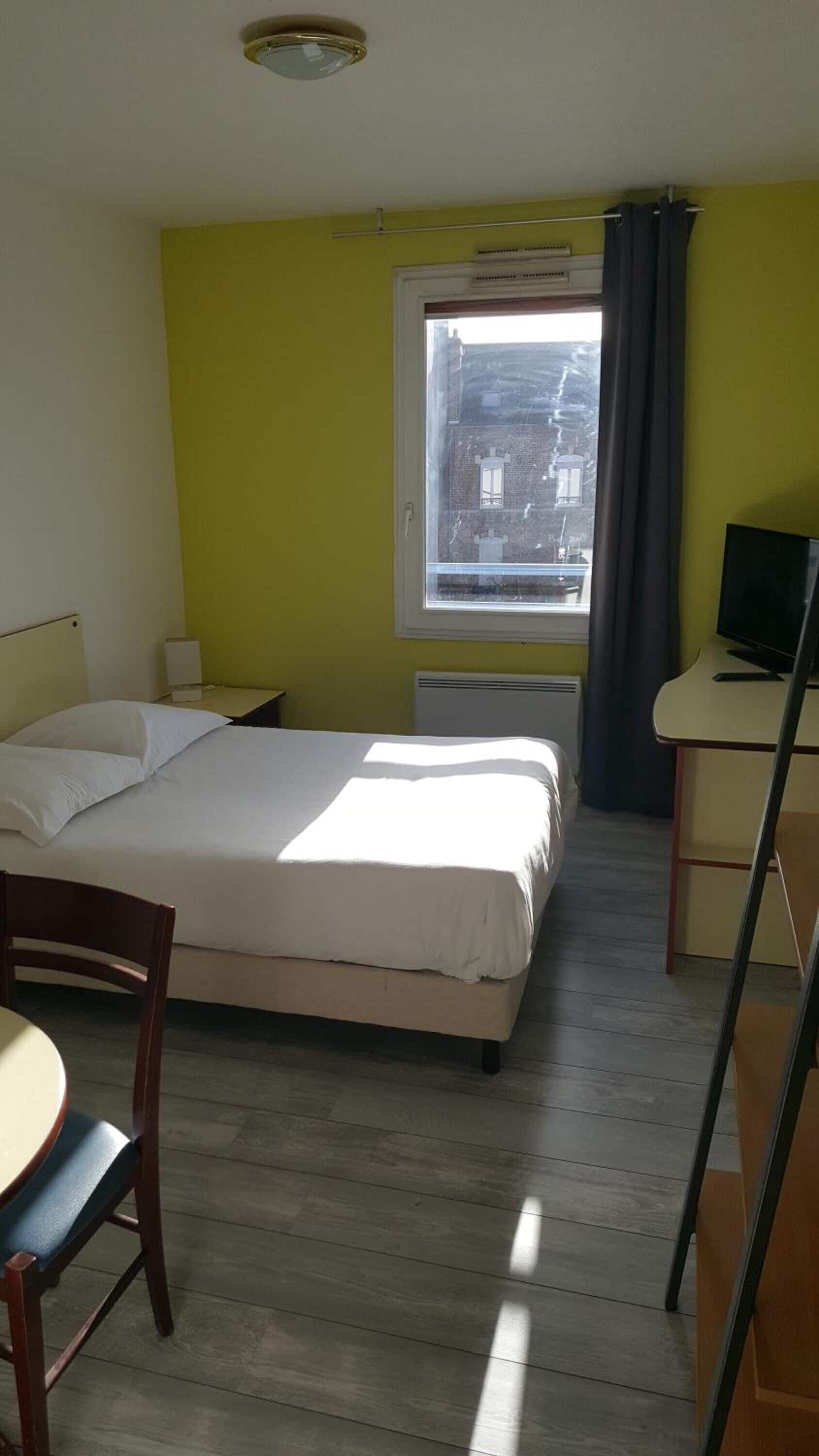 A hotel room with a bed, a desk, a chair, a TV, and a window with a view of buildings.
