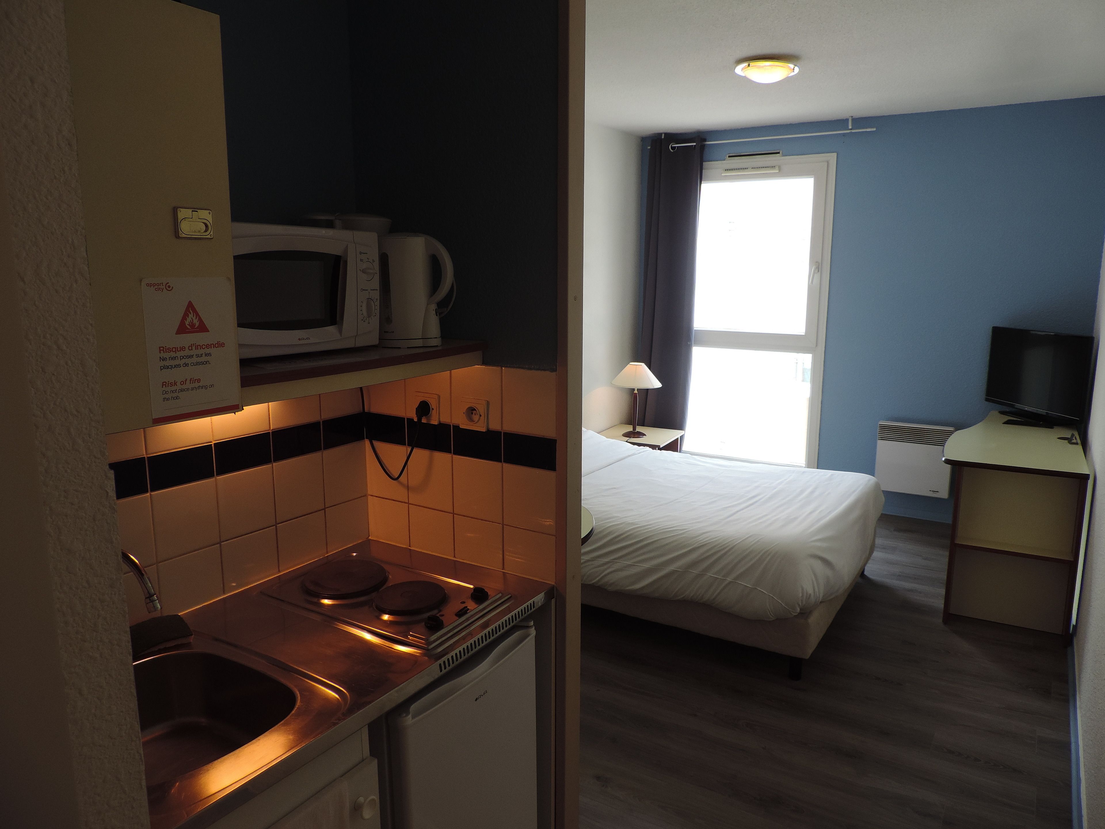 Quarto de hotel com cozinha compacta, uma cama, televisão e janela com cortinas.