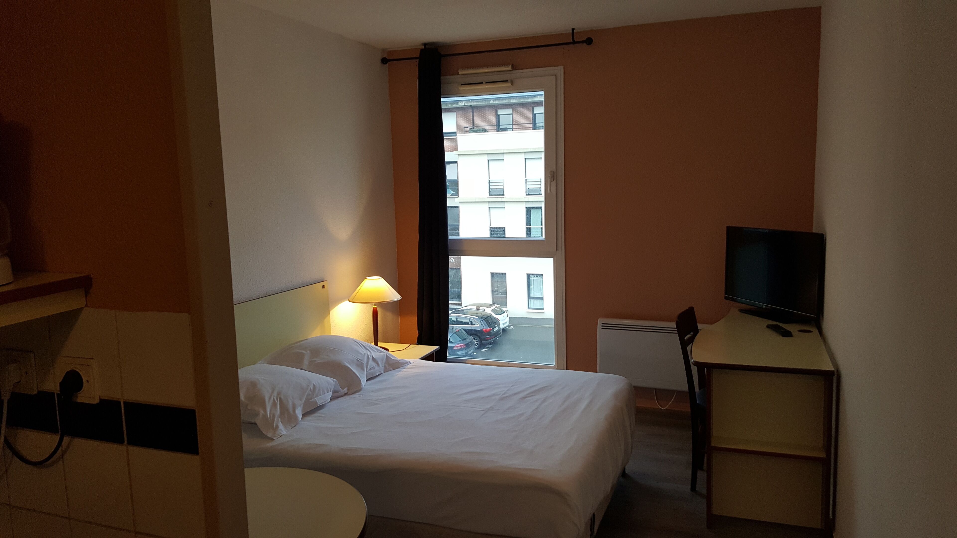 A hotel room with a bed, a desk with a television, a lamp, and a view of a parking area and buildings outside.