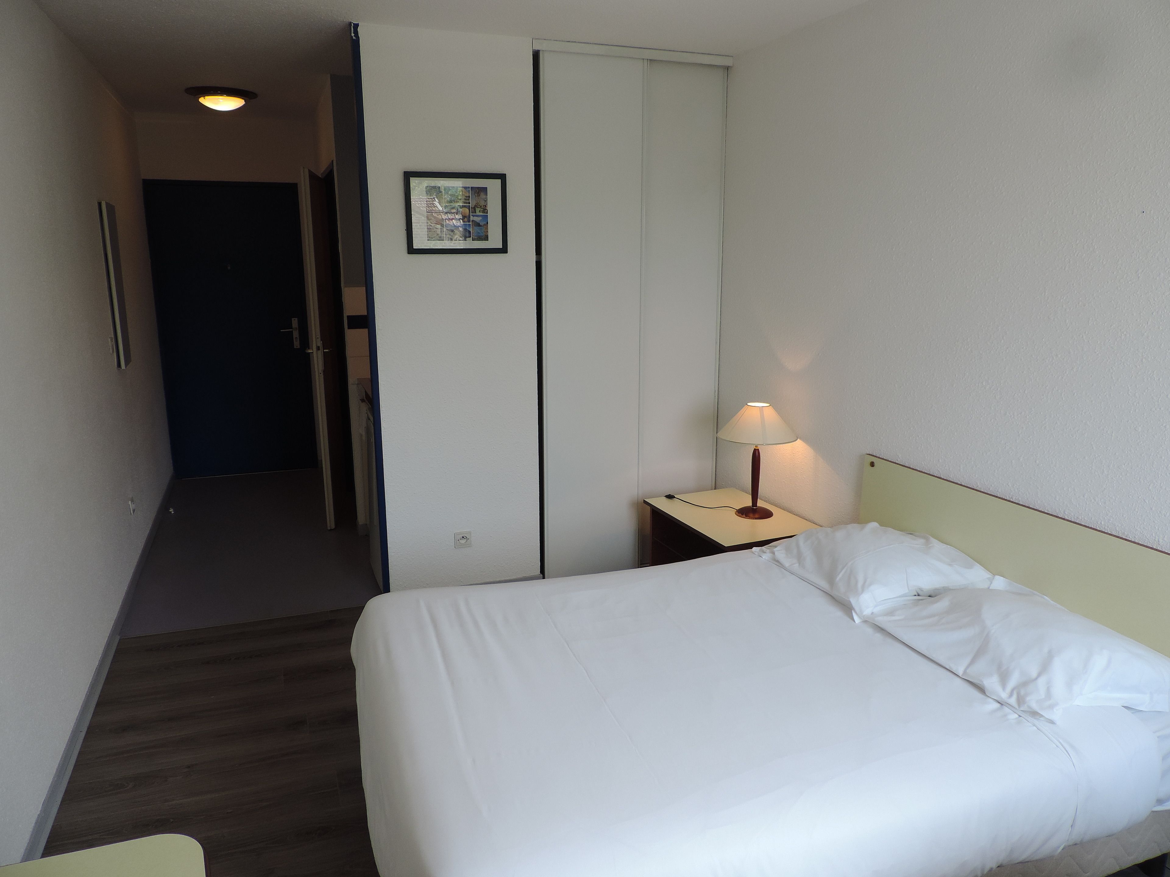Quarto de hotel com cama, mesa de cabeceira com abajur, porta e um quadro na parede.