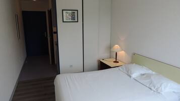 Quarto de hotel com cama, mesa de cabeceira com abajur, porta e um quadro na parede.