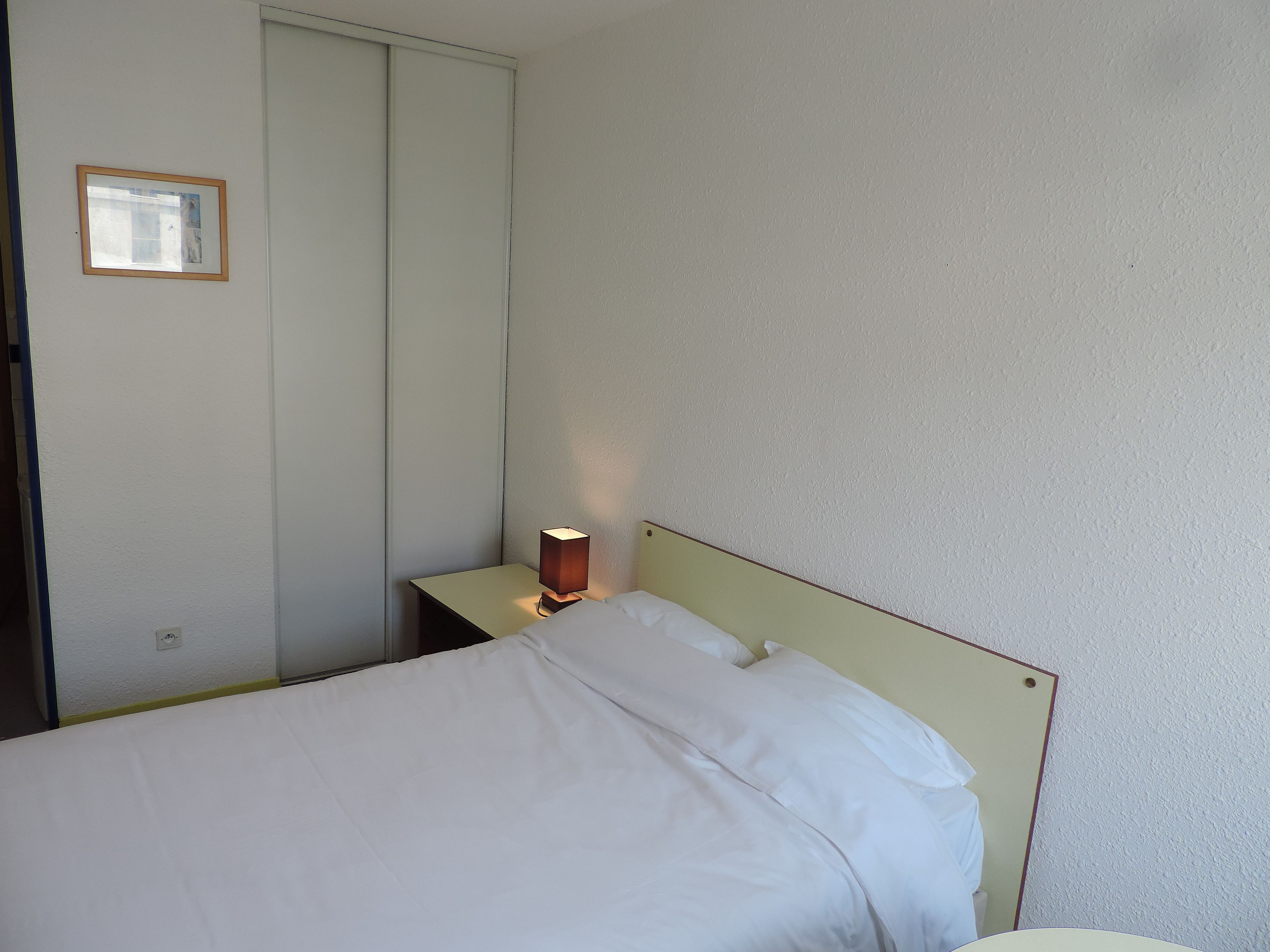 A single bed with a wooden headboard, a bedside table with a lamp, and a wardrobe in a room with plain walls.