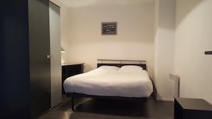 A neatly made bed in a small, dimly lit room with a single nightstand and a framed picture on the wall.
