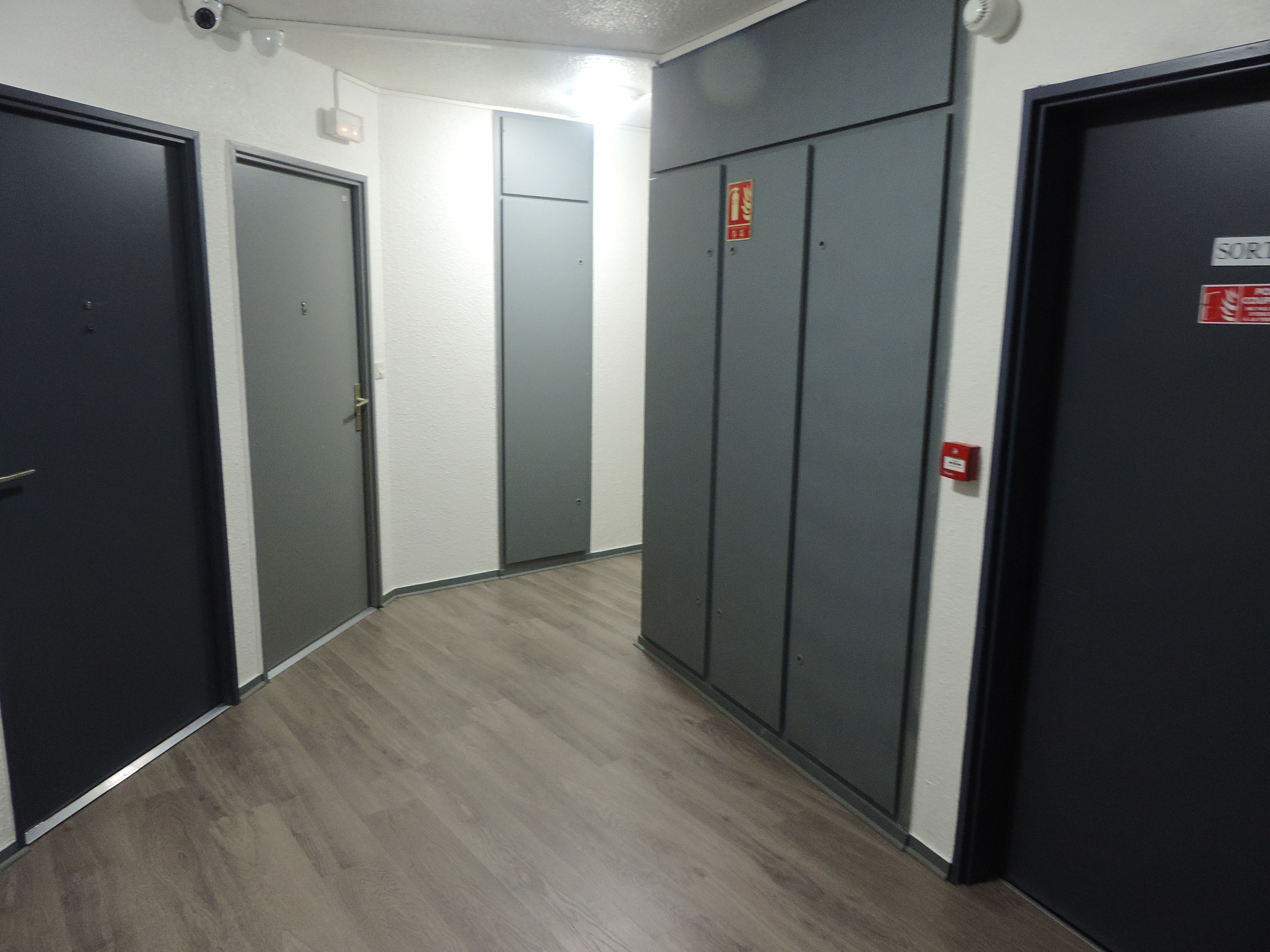 A hallway with grey doors, a fire alarm, and a security camera.