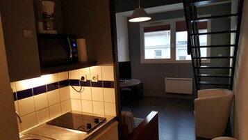 Cozinha compacta com fogão, micro-ondas e painel de azulejos. Há uma janela com persianas e um radiador na parede.