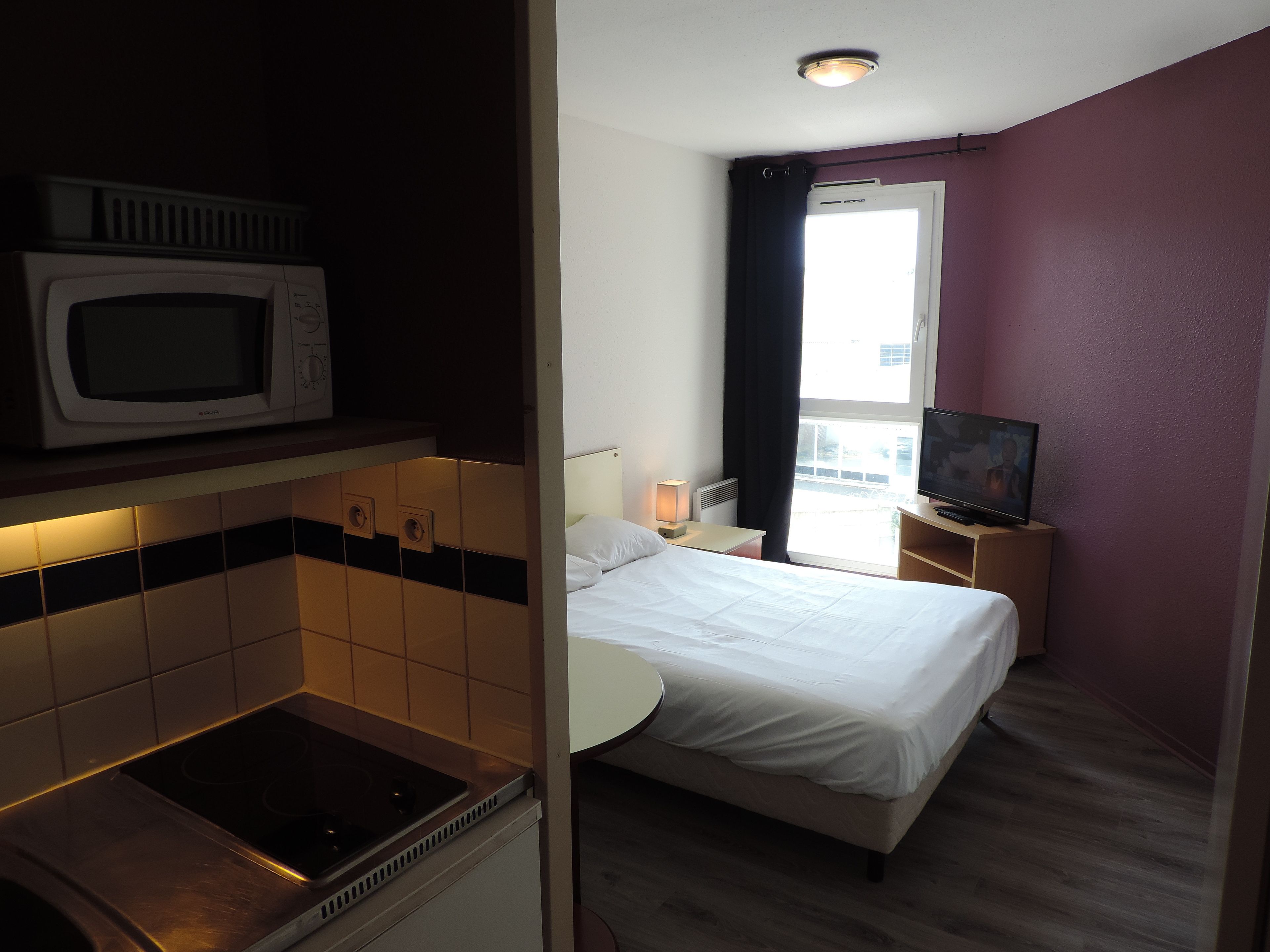 A hotel room with a kitchenette, a bed, a TV, and a window with curtains.