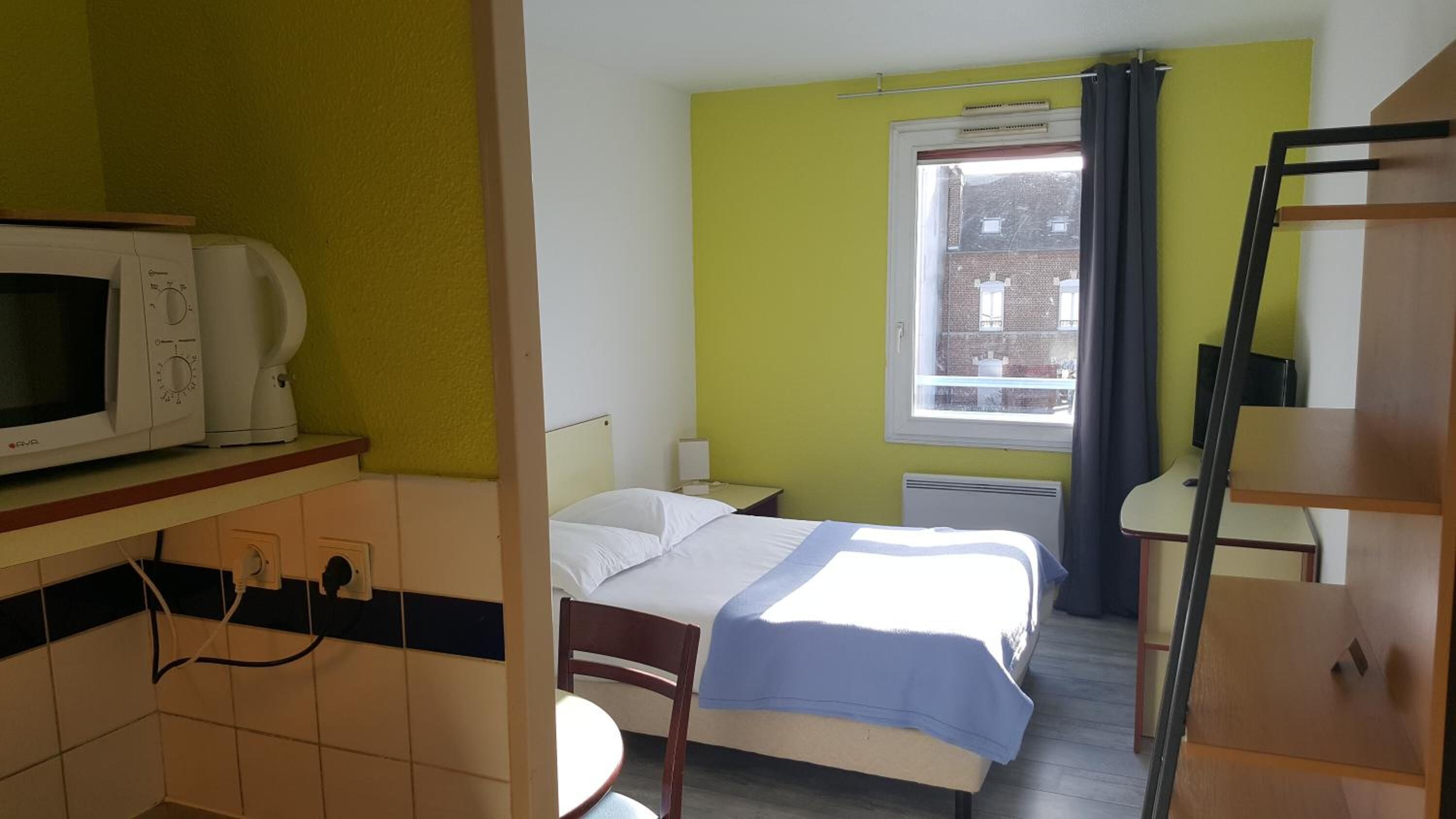 Een kleine, compacte hotelkamer met een bed, nachtkastje, magnetron en raam met gordijnen.
