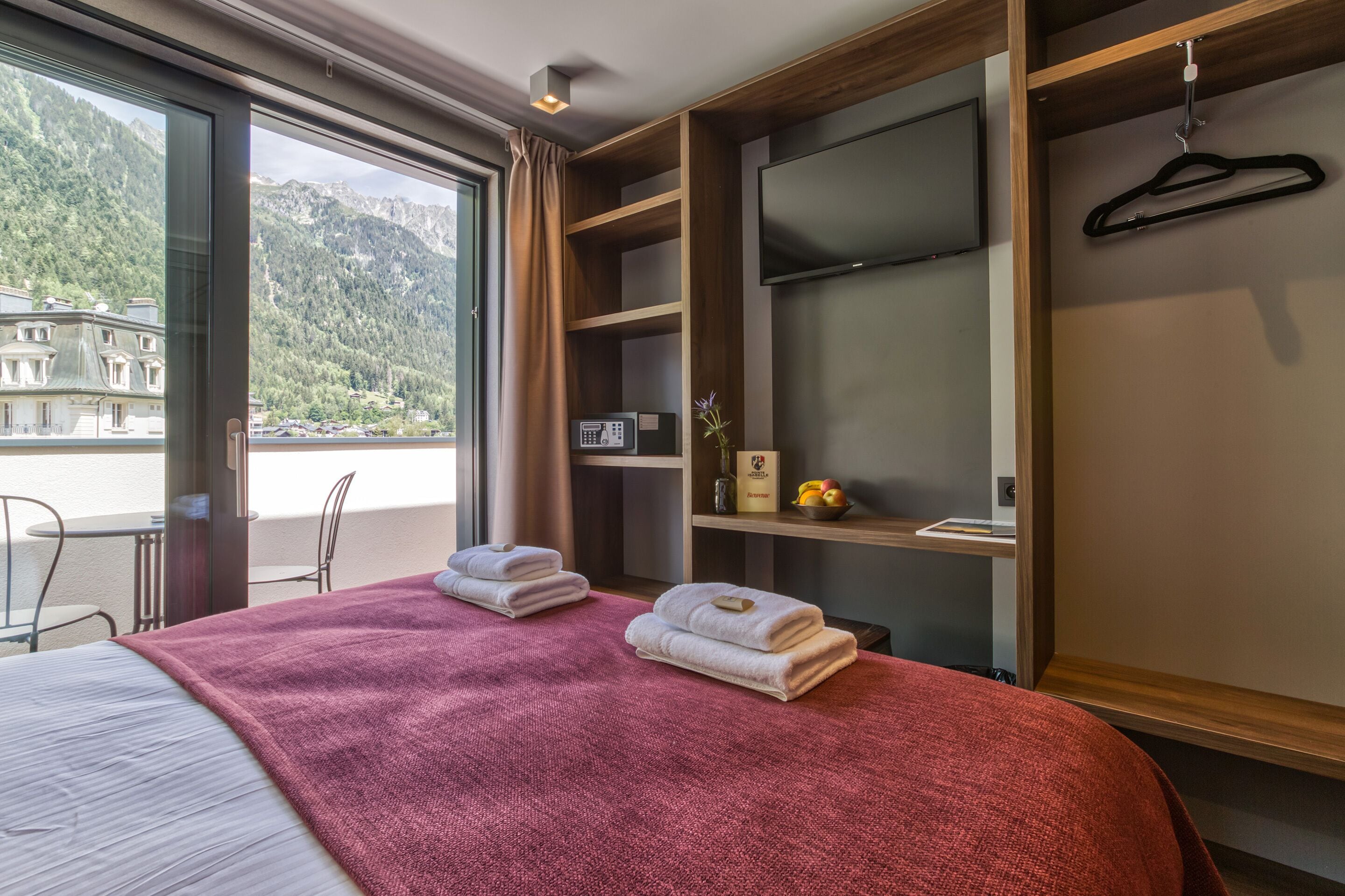 Habitación de hotel con cama, manta roja, televisor, estante de madera y vista a las montañas.