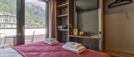 A hotel room with a bed, a red blanket, a TV, a wooden shelf, and a view of mountains.