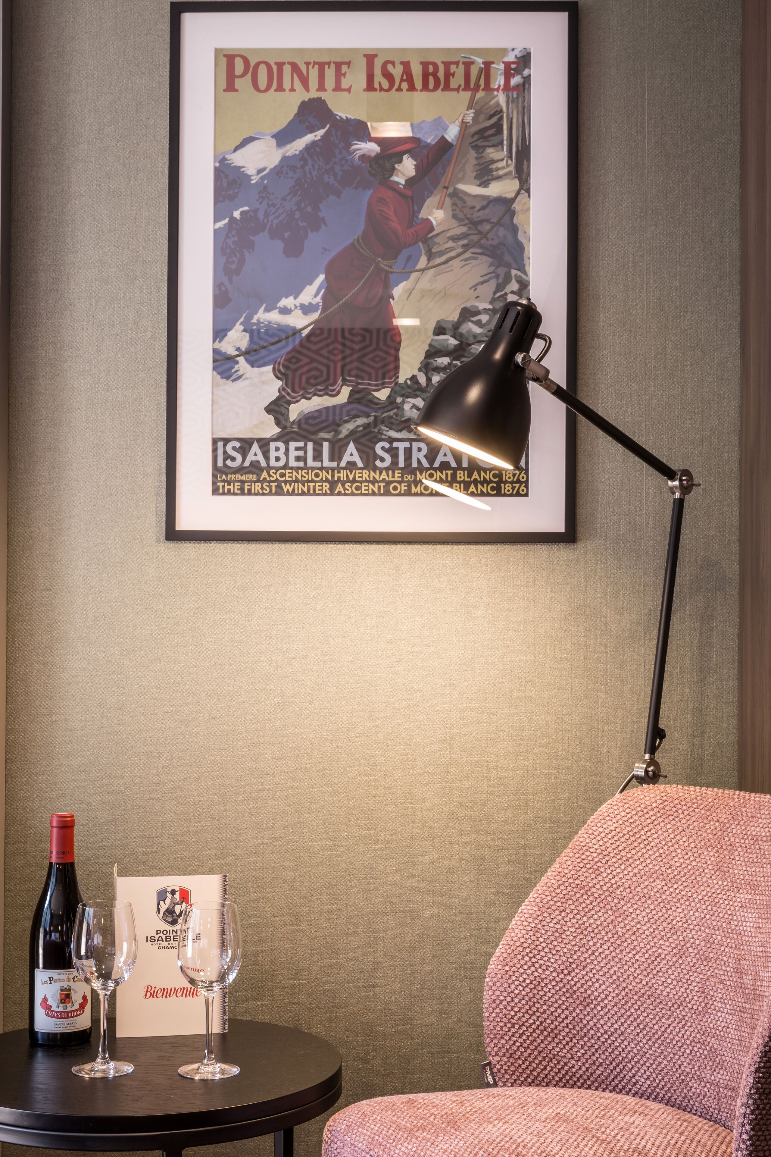 A framed poster of Pointe Isabelle on a wall, a table with a bottle of wine and glasses, and a red upholstered chair.