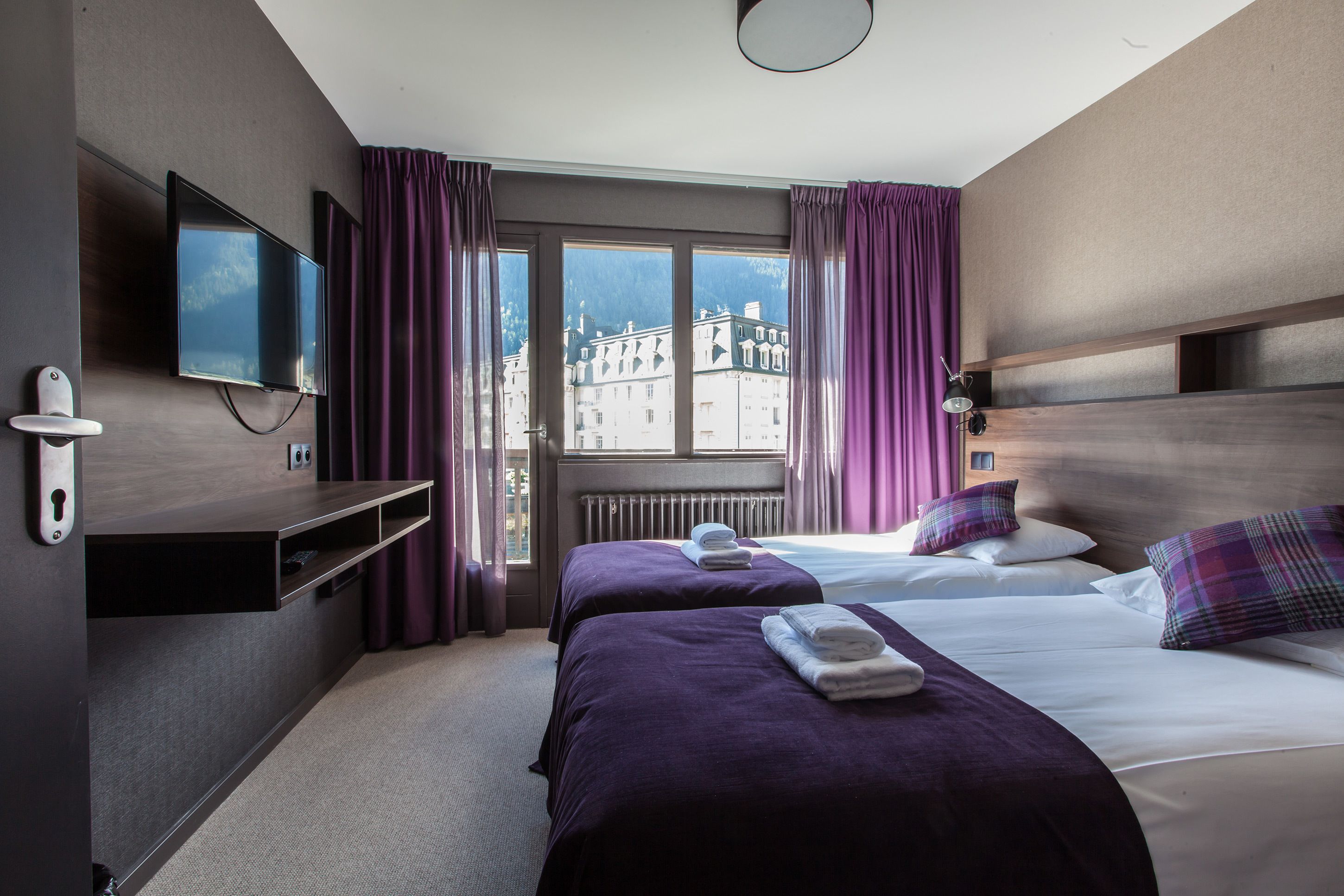 A hotel room with two beds, a TV, a window with curtains, and a view of buildings.