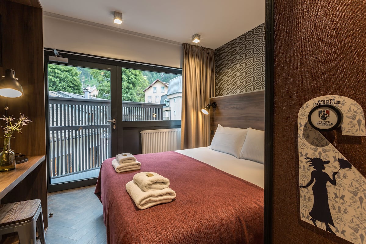 Ett hotellrum med en stor säng, ett skrivbord med en stol, ett fönster med utsikt över hus och en vägg med ett mönster.