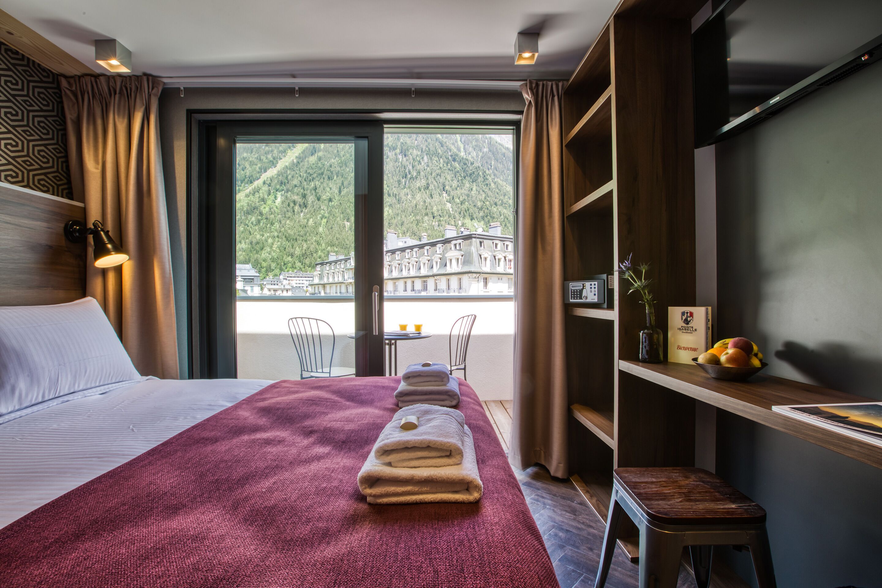 Habitación de hotel con una cama grande, un cabecero de madera, un escritorio con silla, un taburete pequeño, una estantería con un florero y fruta, y vistas a una montaña y edificios a través de un ventanal amplio.
