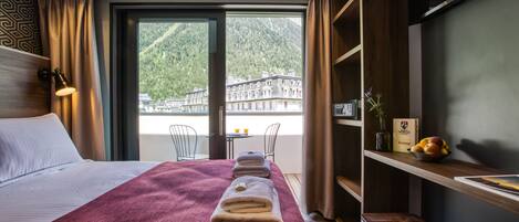 Habitación de hotel con una cama grande, un cabecero de madera, un escritorio con silla, un taburete pequeño, una estantería con un florero y fruta, y vistas a una montaña y edificios a través de un ventanal amplio.