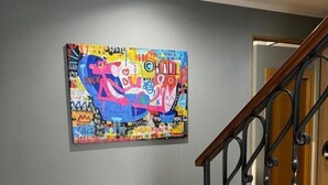 Una escalera con barandilla de madera y balaustradas negras, que conduce a una habitación con pared gris y un cuadro abstracto de colores.