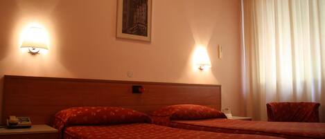 Habitación de hotel con dos camas, cada una con una colcha floral roja y cabecero. Dos lámparas de pared proporcionan luz, y hay un cuadro enmarcado en la pared.