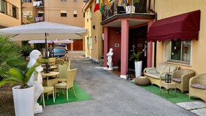 Un espacio al aire libre con mobiliario de mimbre y una estatua, un edificio con balcones y banderas, y un automóvil estacionado.