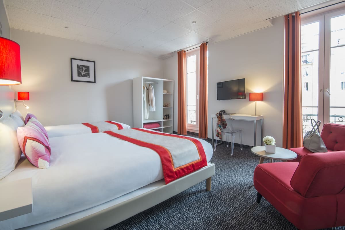 Ein Hotelzimmer mit zwei Betten, einem Schreibtisch, einem Stuhl, einem Fernseher und einem Balkon mit orangenen Vorhängen.