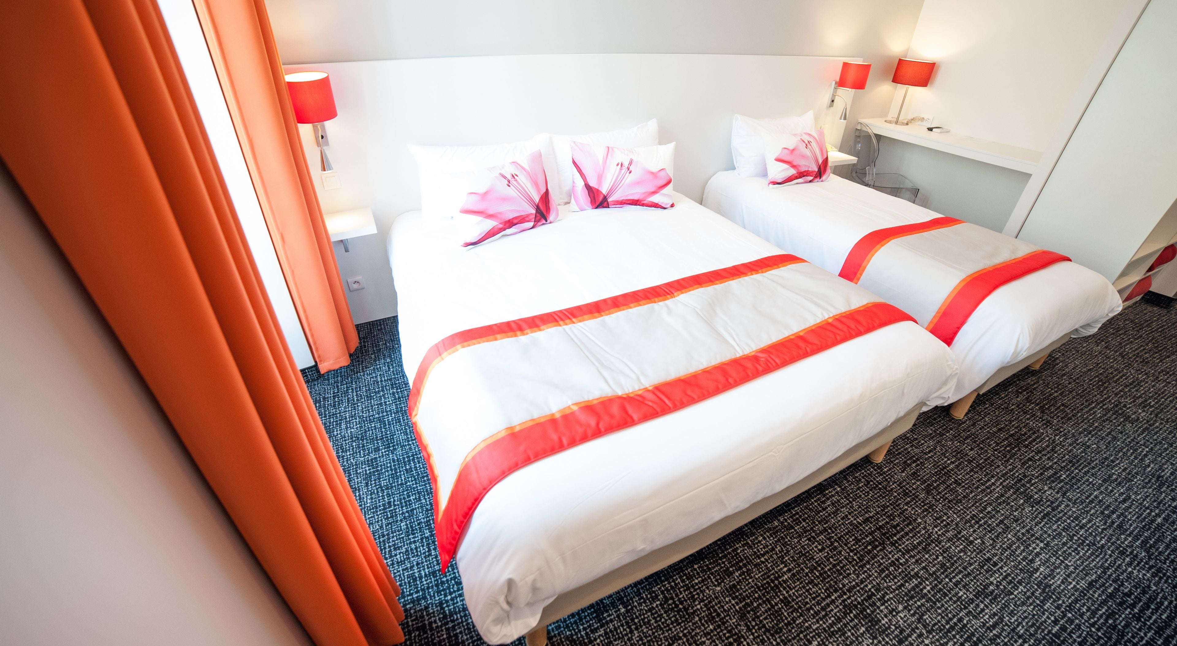 A hotel room with two beds, each with white bedding and red accents, a small desk with a lamp, and a window with curtains.