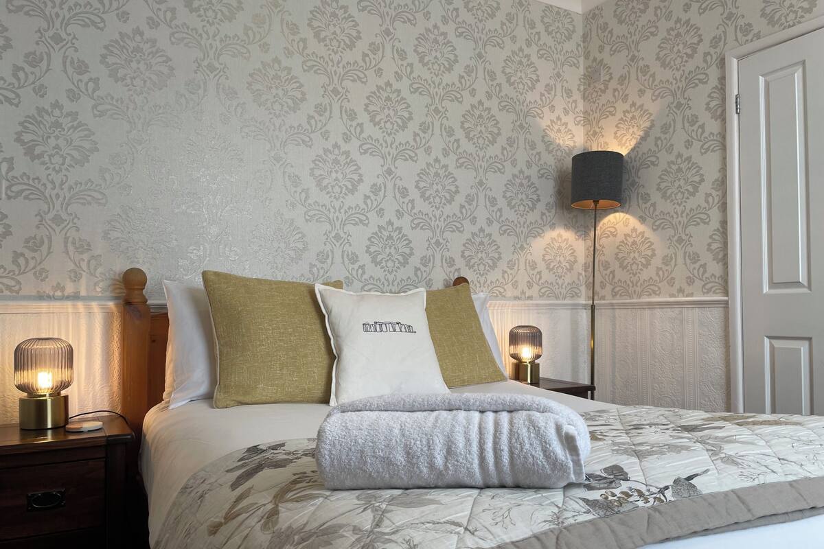 Ein ordentlich bezogenes Bett mit Blumenmuster, zwei Nachttische mit Lampen und eine Wand mit ornamentalem Wandpapier.