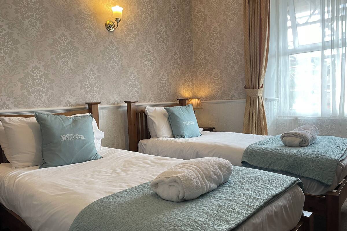 Ein Hotelzimmer mit zwei Betten, jedes mit einem blauen Kopfkissen und einer weißen Decke. Über den Betten befindet sich eine Wandleuchte, und es gibt ein Fenster mit Vorhängen.