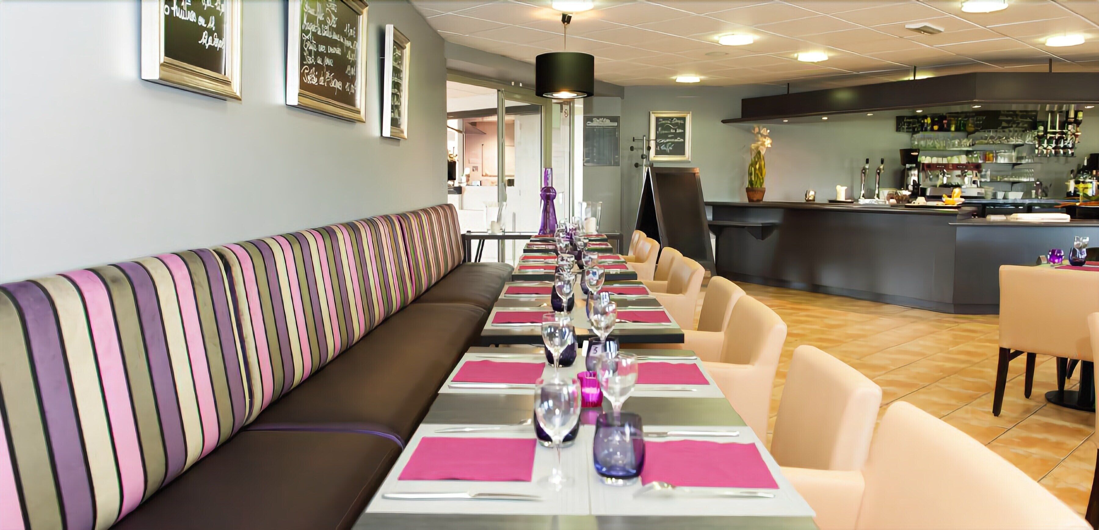 A restaurant with a long dining table set with pink napkins, silverware, and wine glasses. There is a striped bench along the wall and a bar area in the background.